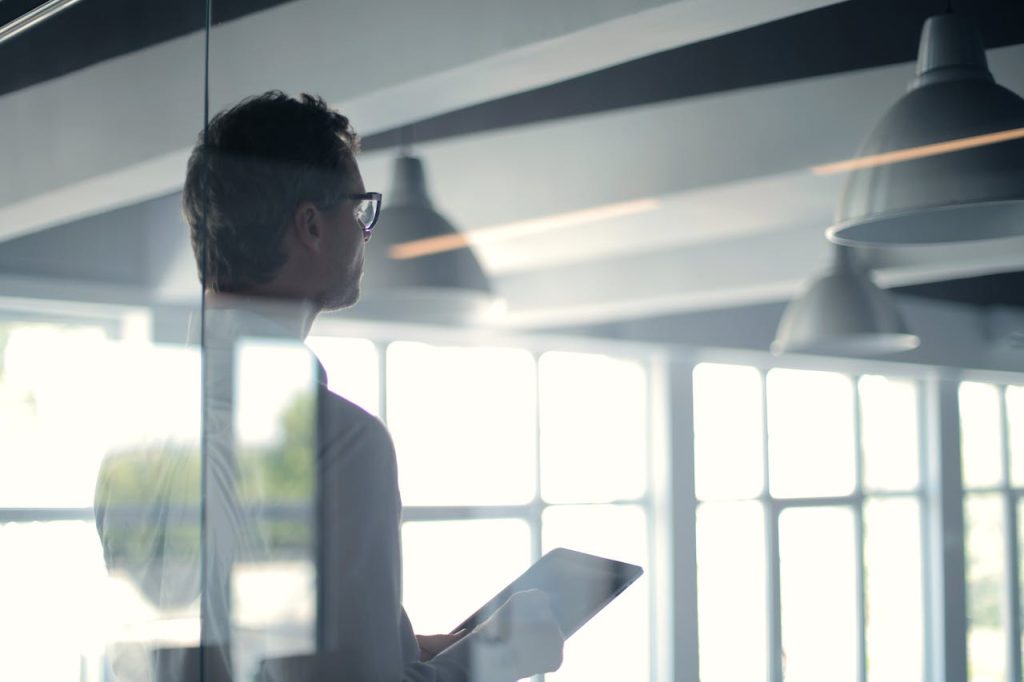 formal-man-with-tablet-giving-presentation-in-office-3760093 A businessman holding a tablet in an office, looking thoughtfully through a glass wall.
