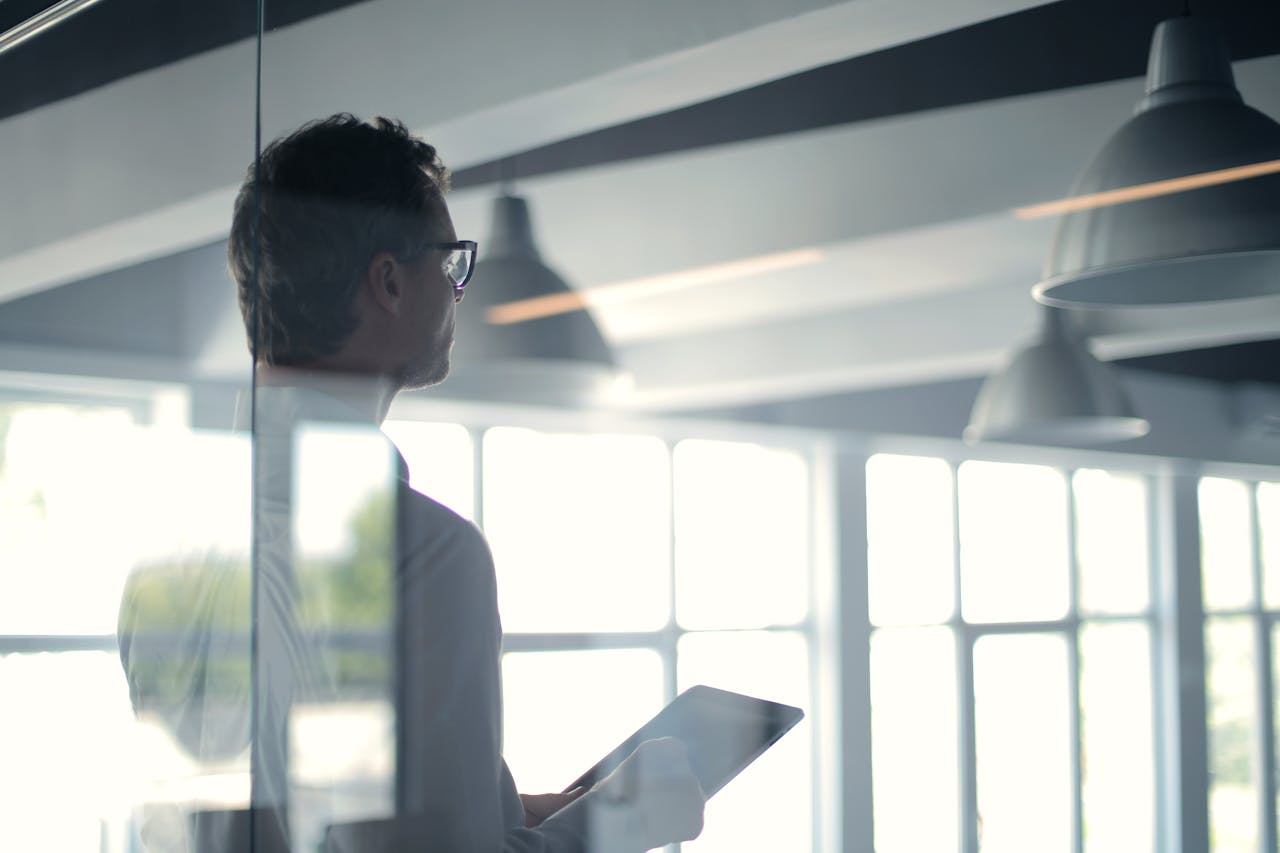 services-04 A businessman holding a tablet in an office, looking thoughtfully through a glass wall.