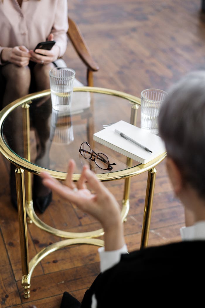 about-img-01 Two adults engaged in a therapy session, seated at a glass table with notebooks and water glasses.